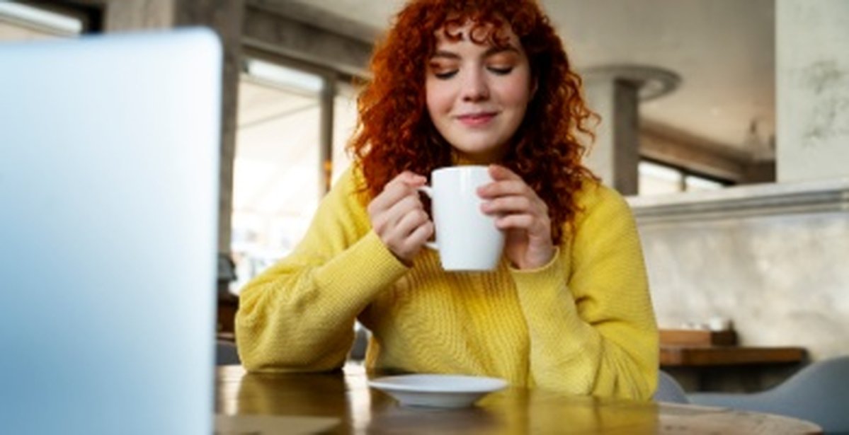 Eine Frau mit roten Haaren und einem Geben Pullover sitzt am Tisch und hat eine Tasse in den Händen und einen Teller vor ihr auf dem Tisch
